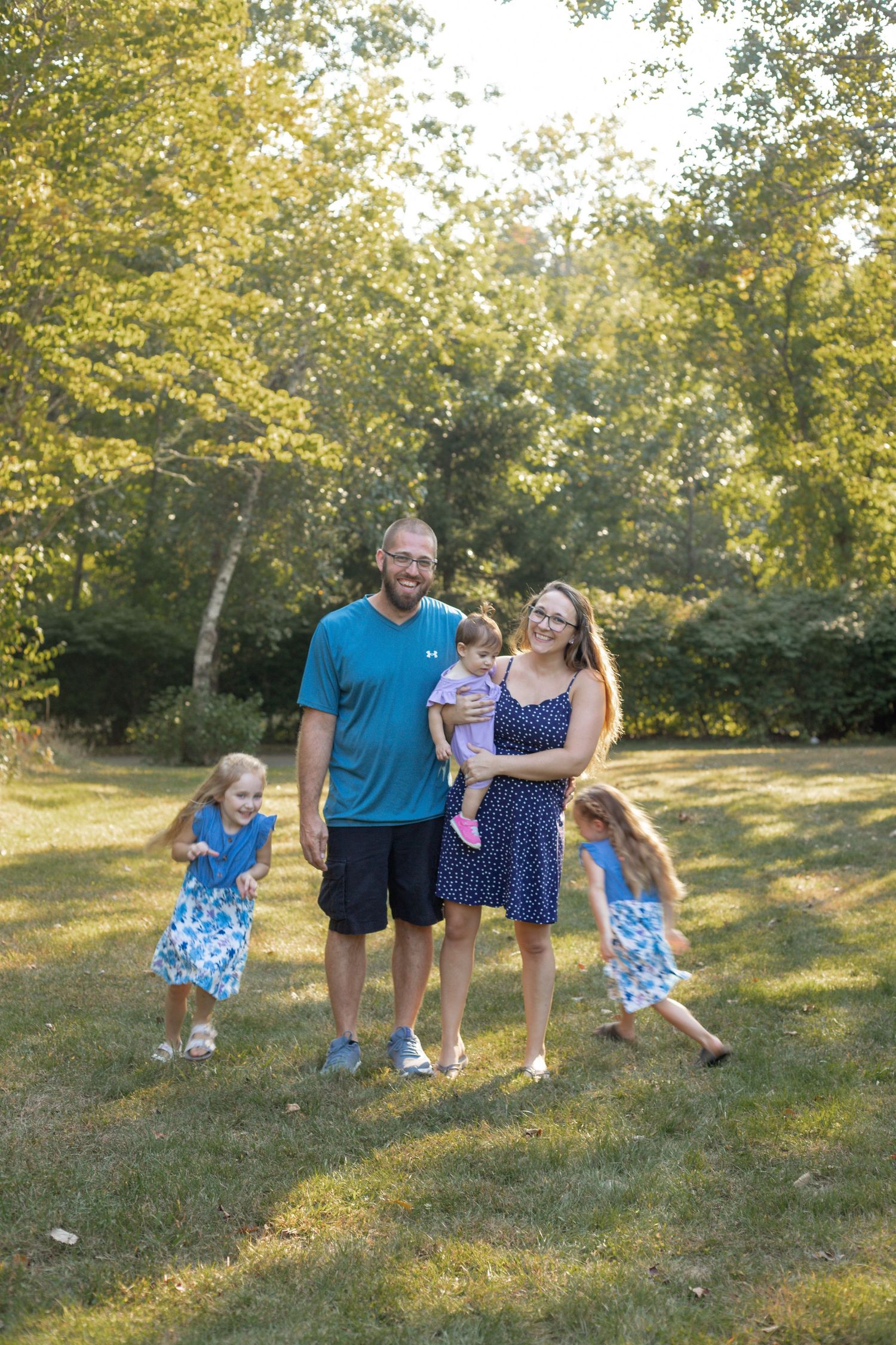 family smiling at the park while the children run circles around mom and dad