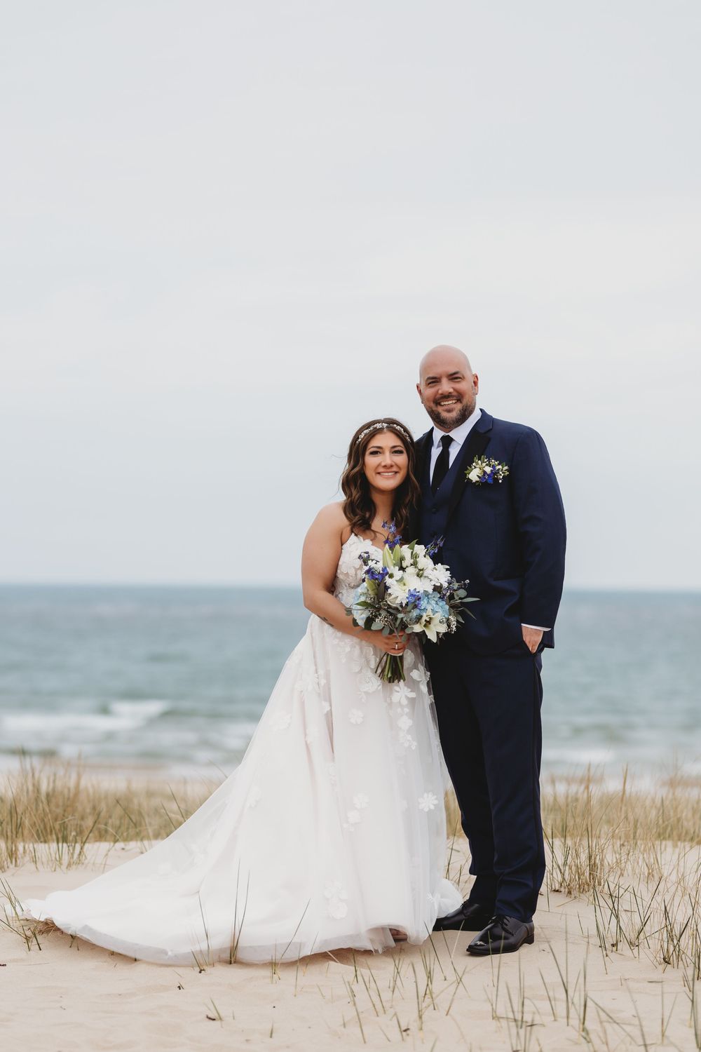 A joyful couple in wedding attire poses together on a sandy beach with ocean and dunes in the background.