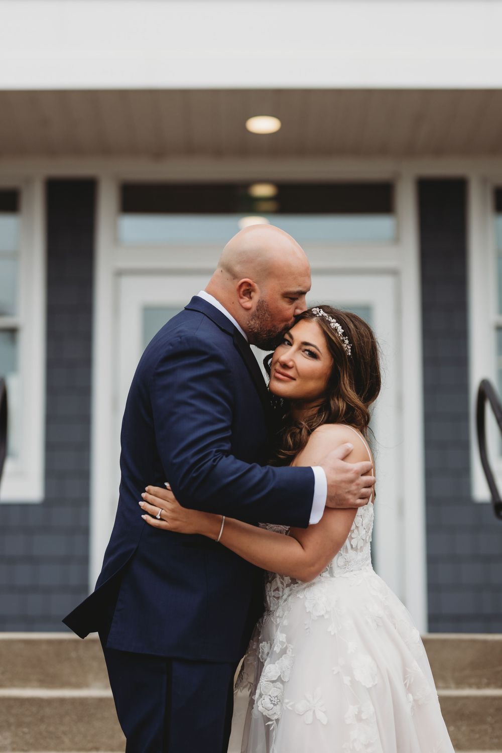 A couple share intimate moments in their wedding attire against a modern house exterior with gray siding.