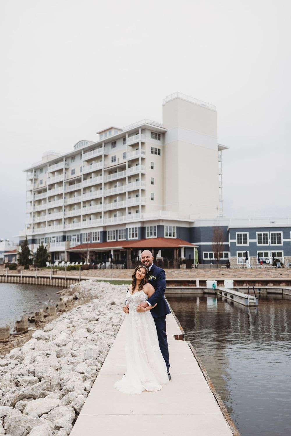 A couple in wedding attire poses on a concrete jetty beside a waterfront hotel building on a cloudy day.