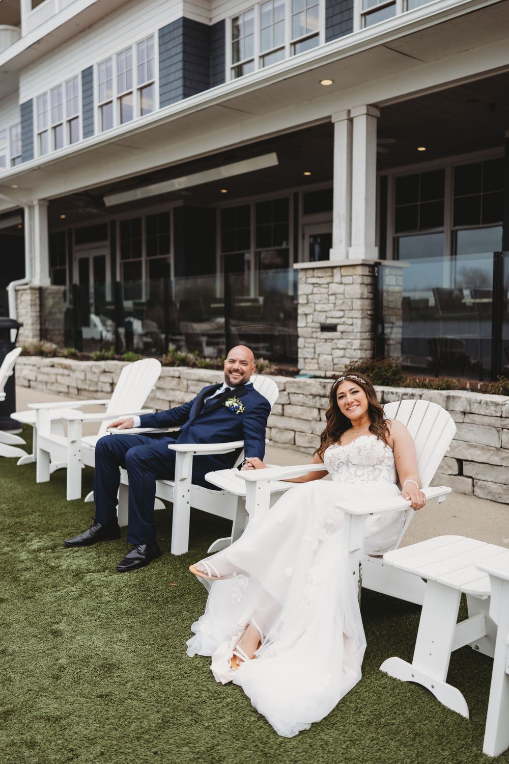 A couple in wedding attire relaxes casually on white Adirondack chairs outside a stone-facade building.