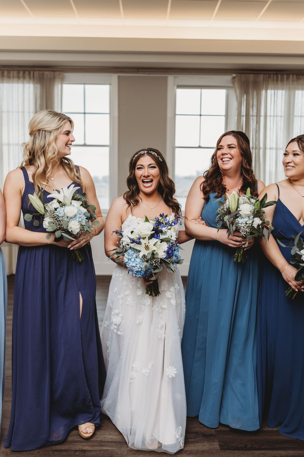 Bridesmaids wearing different shades of blue dresses stand together holding white and blue floral bouquets at a wedding.