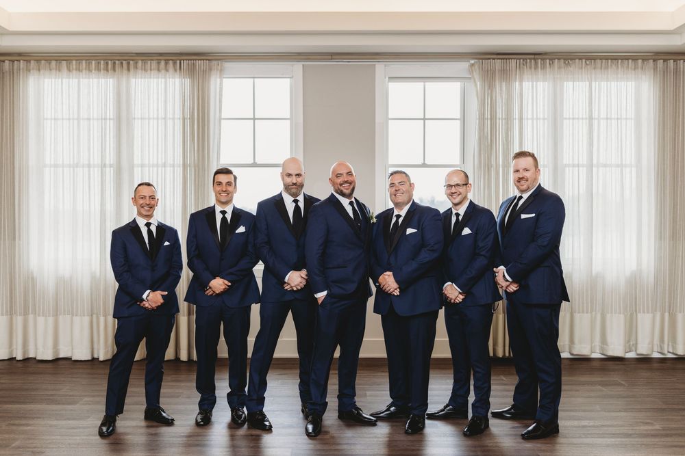 A group of men in matching navy suits stand together in a bright indoor room with white curtains and wood flooring.