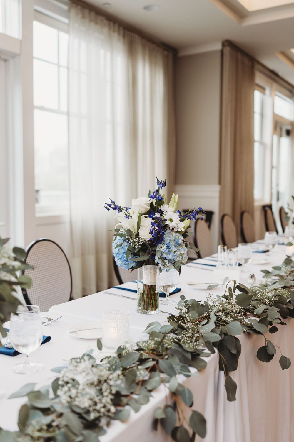 A wedding reception table setting decorated with elegant eucalyptus garland, white linens and blue floral accents.