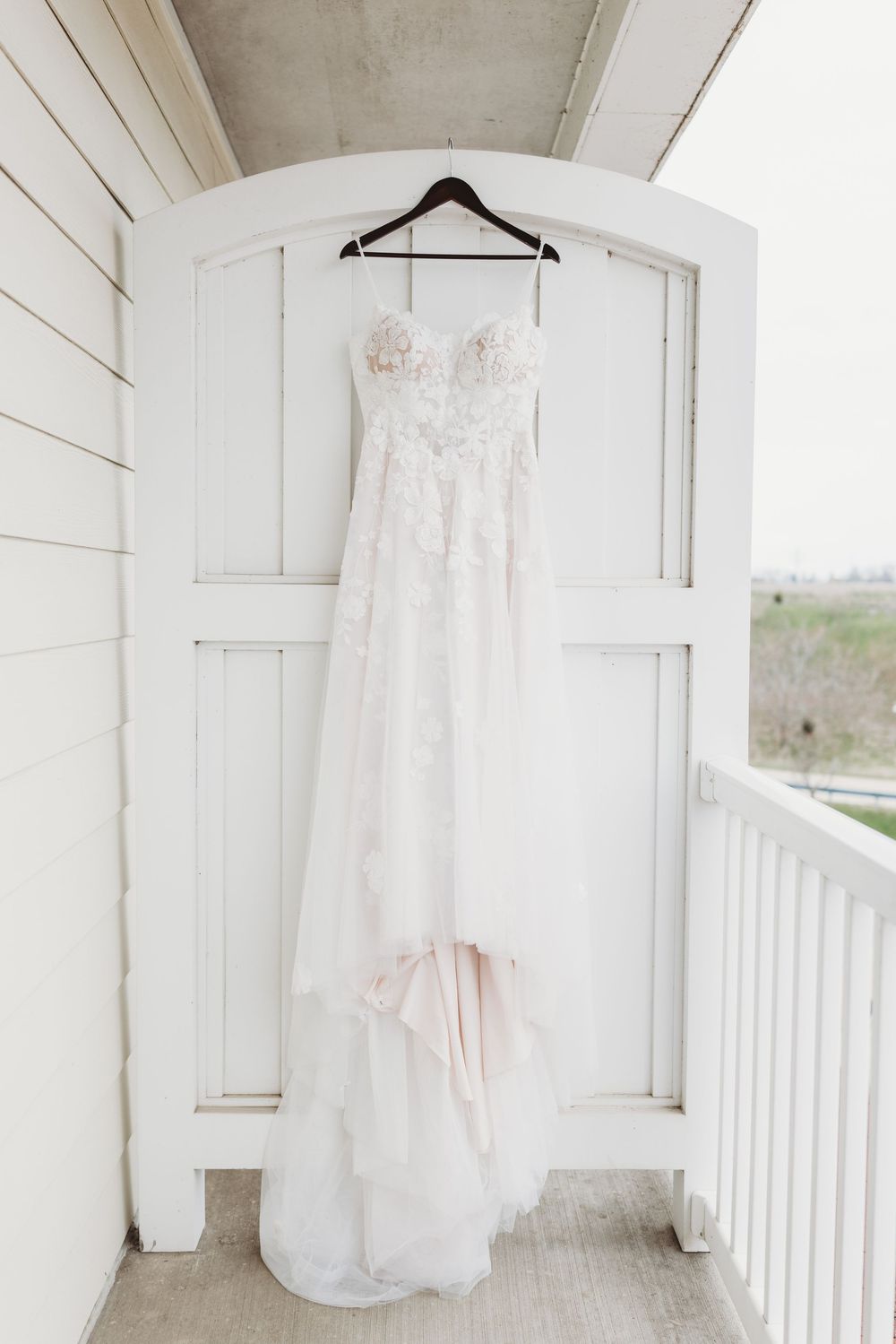 An elegant white lace wedding dress hangs from a hanger on a white door frame of a rustic porch.