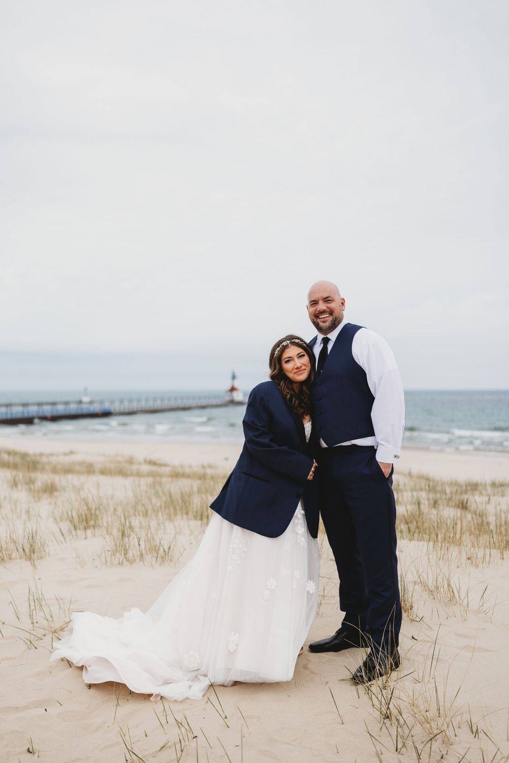 Romantic beach wedding photos captured on a cloudy day with sand dunes and pier in the background.