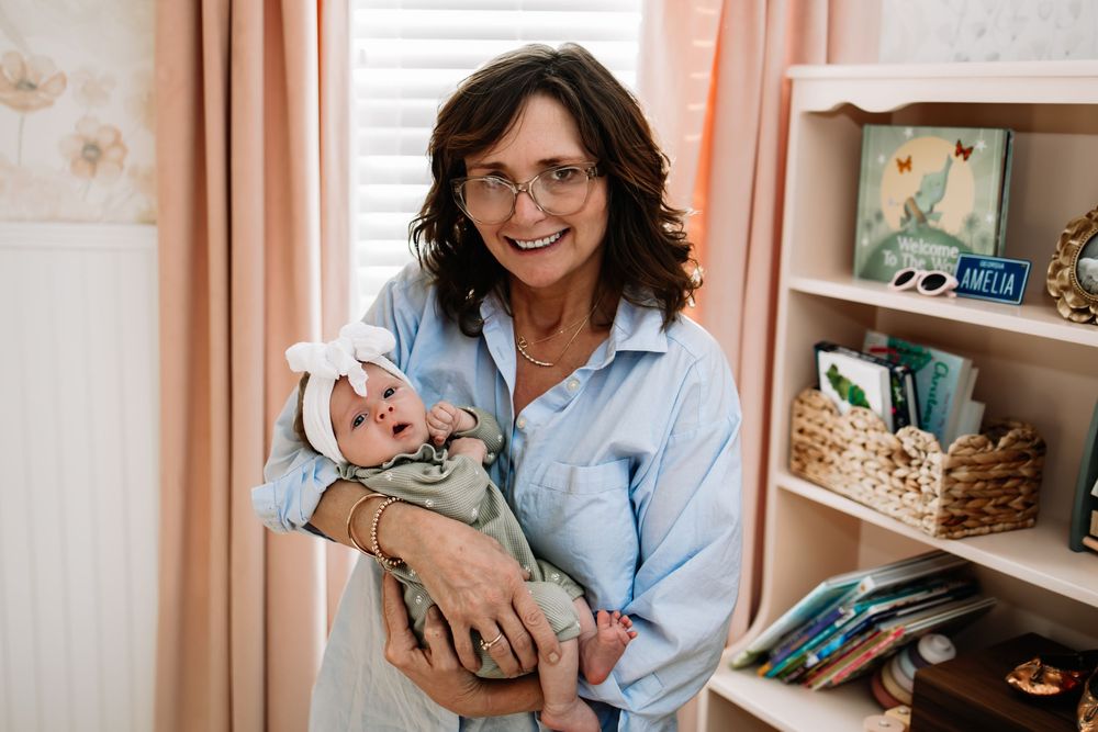 Person in light blue shirt rocking a baby while sitting in a nursery with bookshelves and decor in warm lighting.