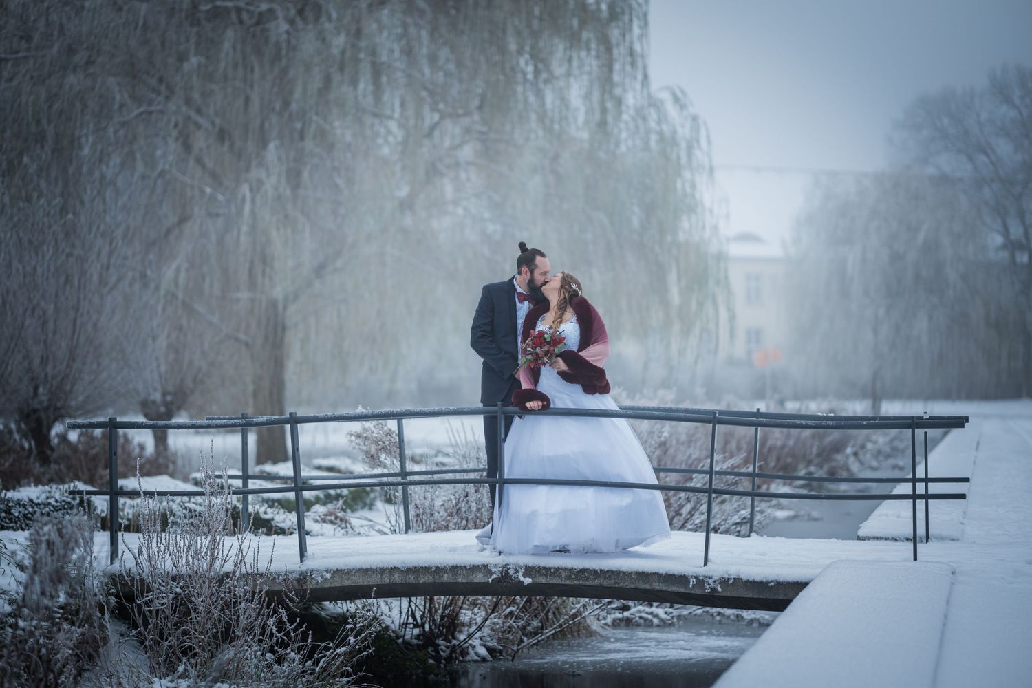 Ein Brautpaar steht auf einer schneebedeckten Brücke im Winter, umgeben von Weiden und Nebel, der eine romantische Atmosphäre schafft.