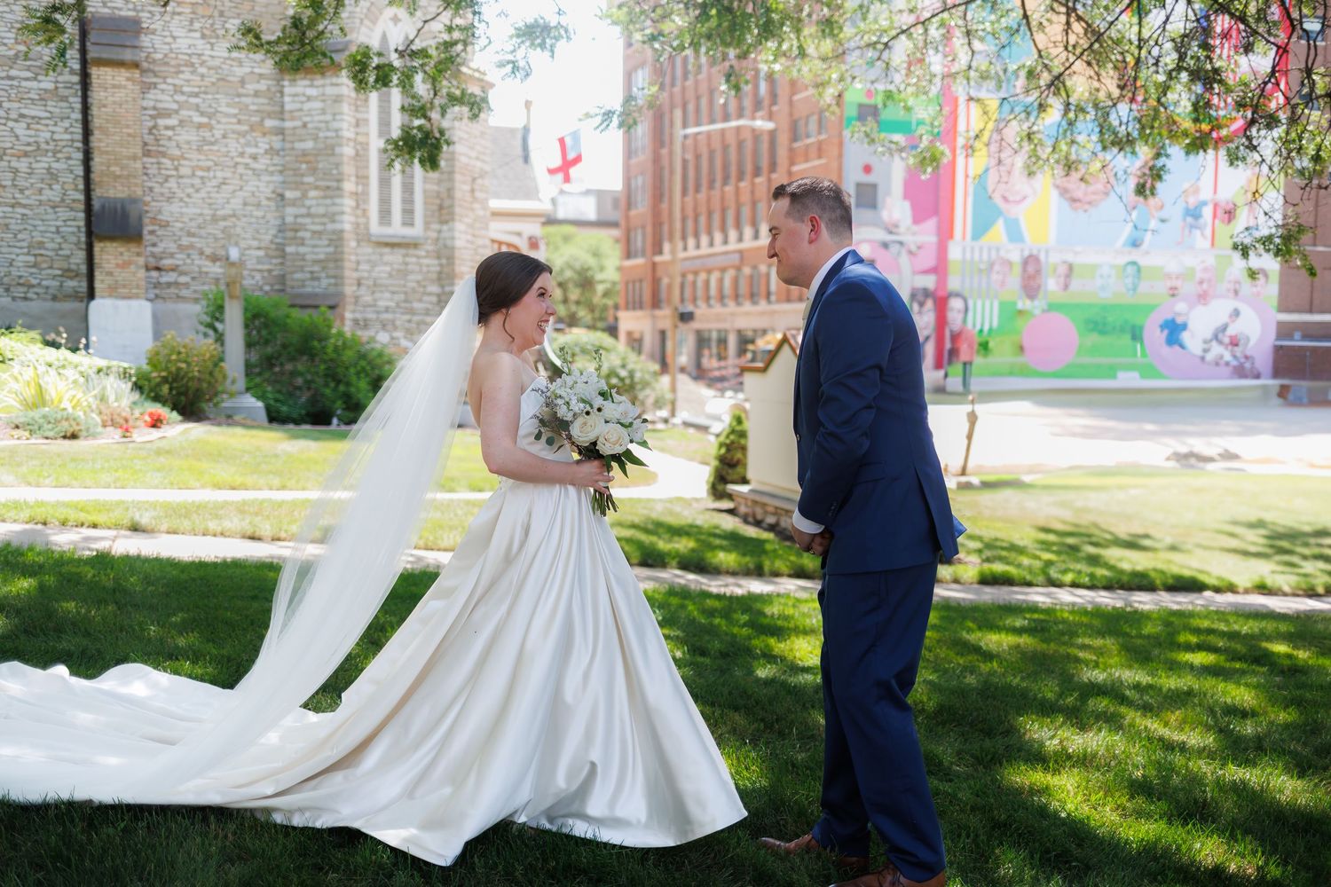 A bride and groom meet for their first look near a brick building surrounded by lush grass and colorful flowers in a garden setting.