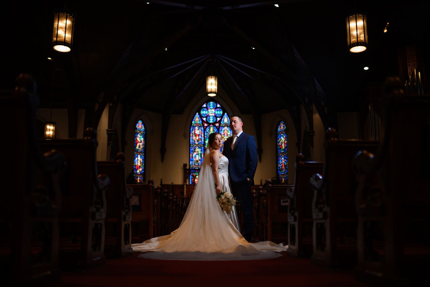 A dramatic wedding ceremony silhouette inside a gothic church with illuminated stained glass windows and warm pendant lighting.