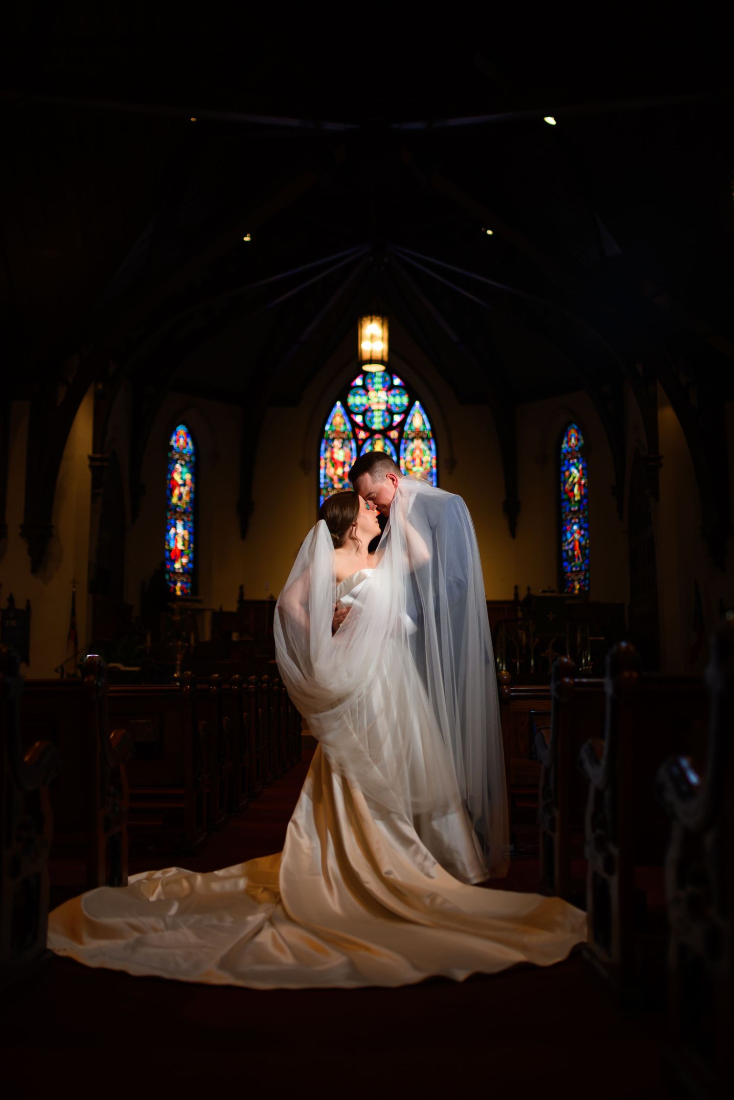 A couple in wedding attire kneels in prayer at a church altar with stained glass windows glowing in the background.