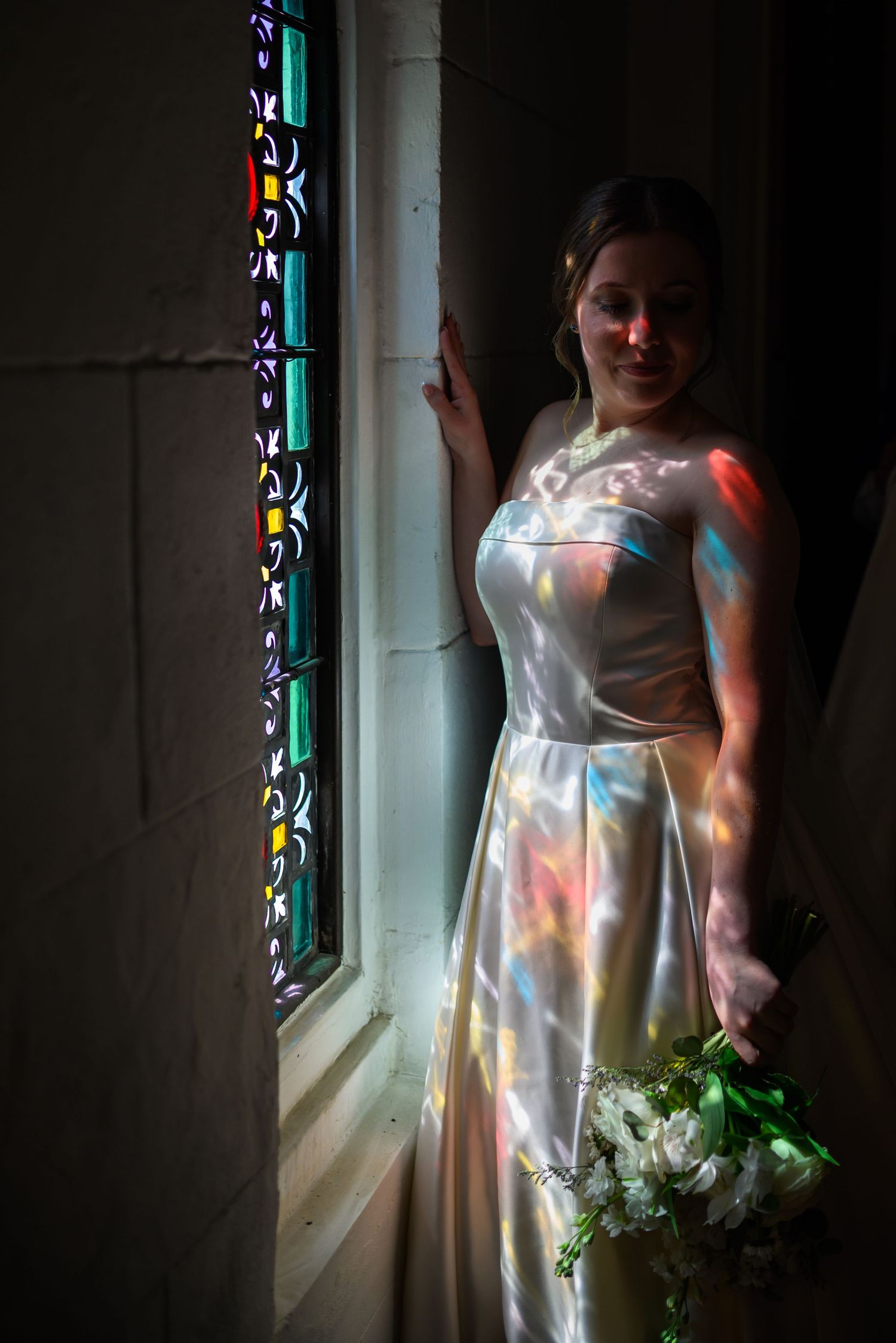 A figure in a white dress stands by a stained glass window while holding a bouquet of flowers in soft lighting.
