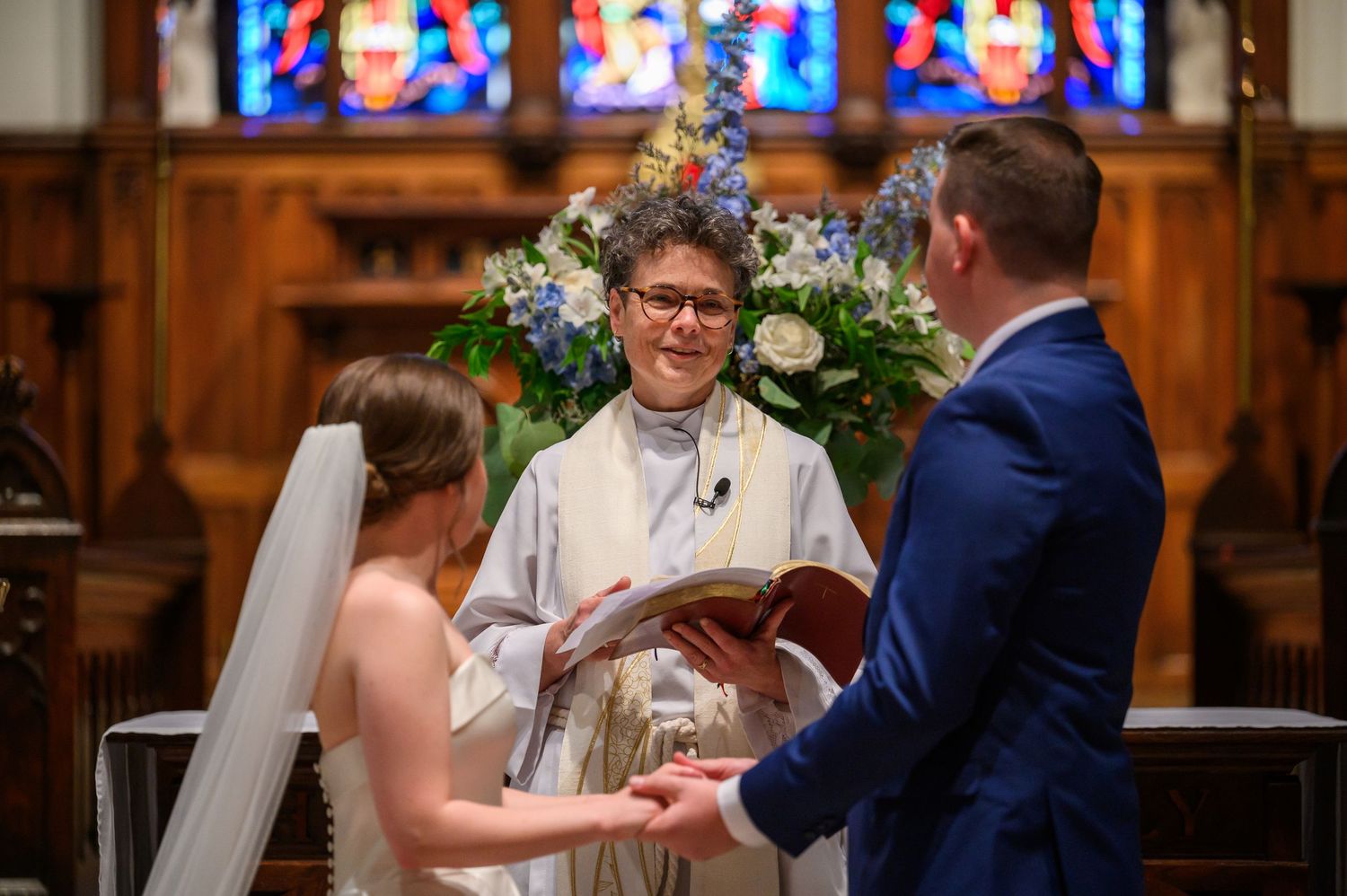 A wedding ceremony takes place inside a church with stained glass windows and floral arrangements.
