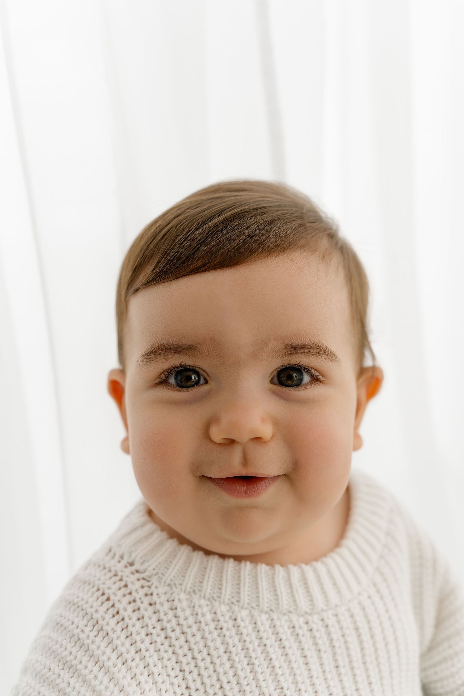 A happy baby wearing a cozy white knit sweater smiles while looking at the camera against a bright background.