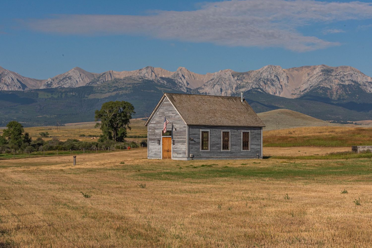 A rustic farmhouse stands solitary against rolling prairie fields with distant mountain peaks in Montana's countryside.