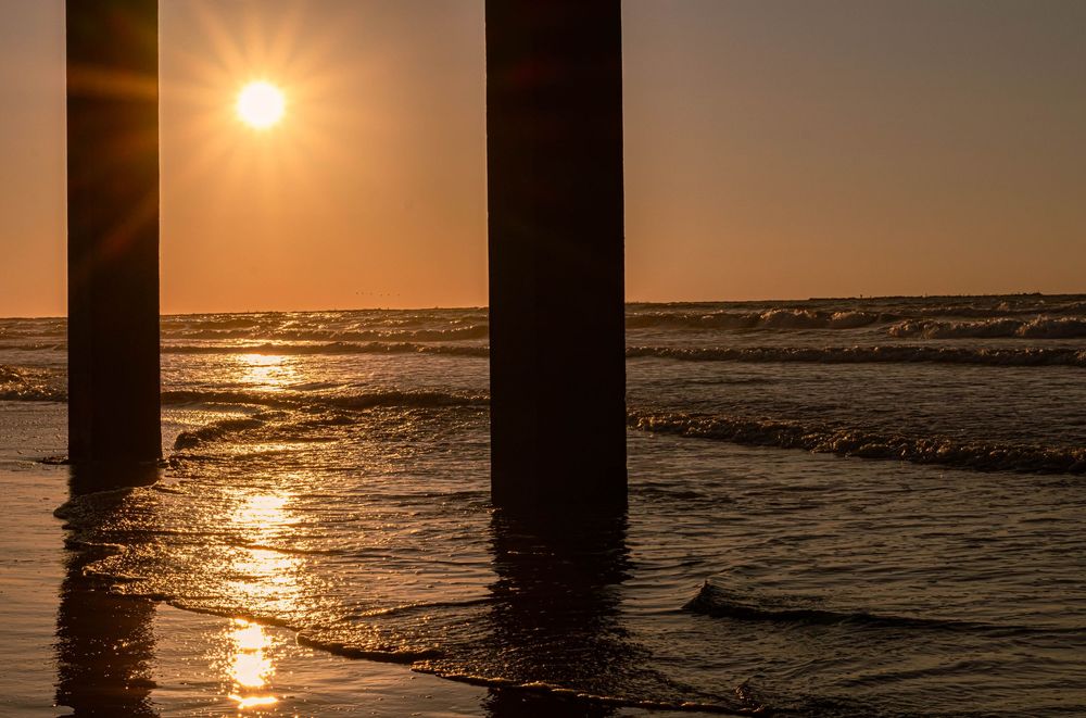 Golden sun setting over ocean waves with dark pier pillars silhouetted against the glowing orange sky and water.