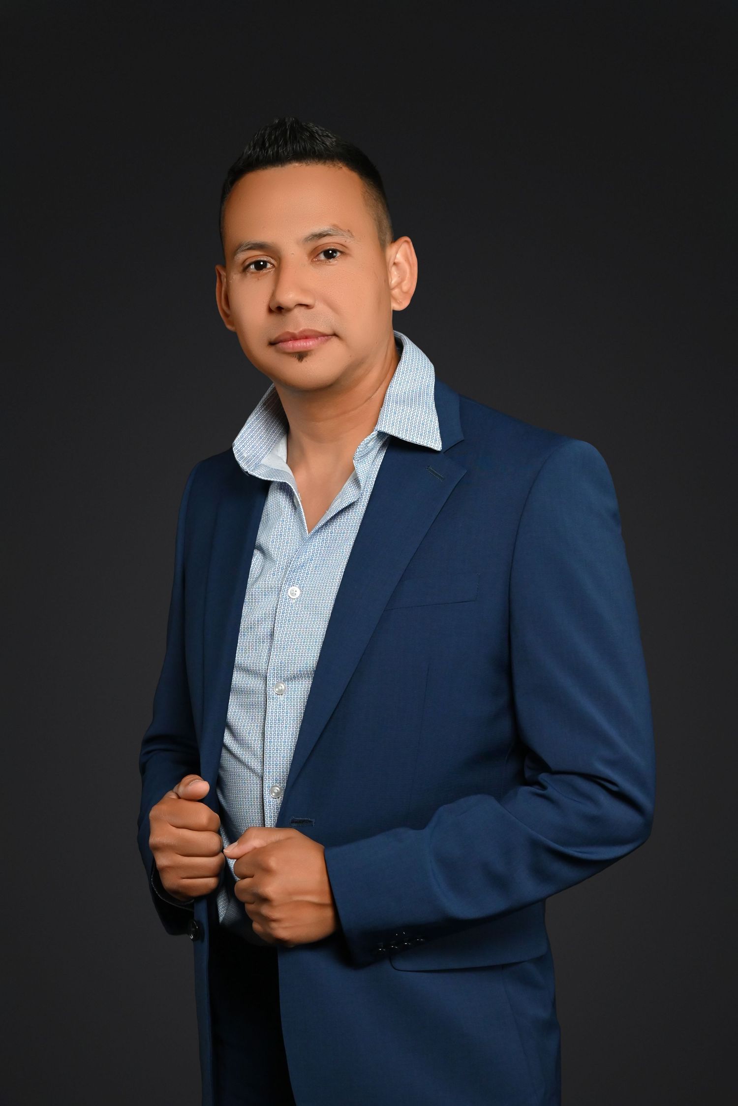 Professional headshot portrait of someone in a navy blue blazer and light blue dress shirt against dark background.