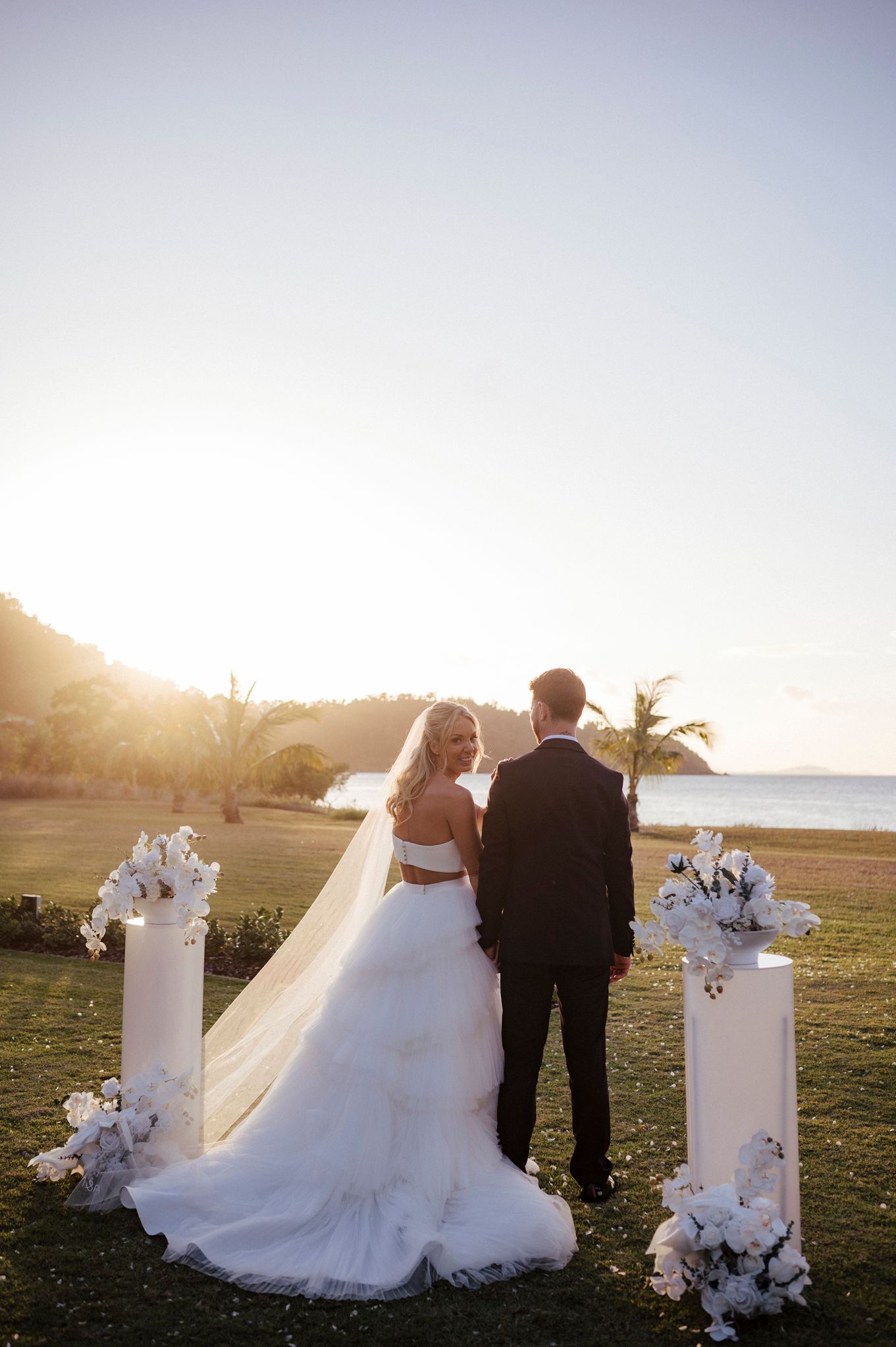 Wedding couple stands between floral pillars at sunset on a beachfront lawn.