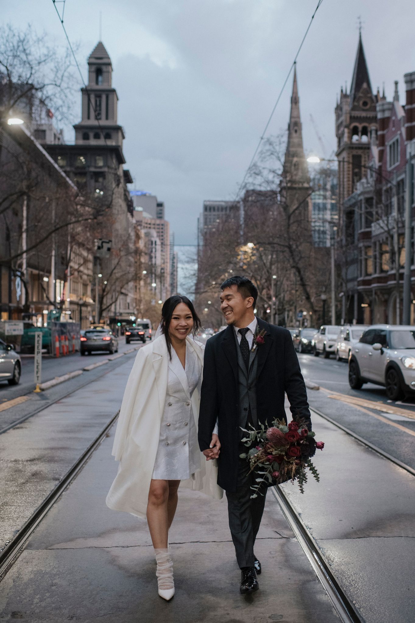 Urban wedding portrait on a rainy city street with gothic architecture and tram lines.