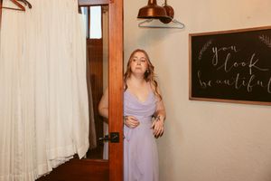 Bridesmaid in lavender dress admires wedding gown hanging in preparation room.