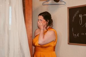 Wedding party member in orange dress has emotional reaction while looking at bridal gown.