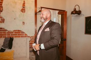 Man in formal grey suit adjusting his tie in a rustic brick-walled room.