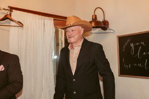 A person wearing a brown cowboy hat and black blazer stands indoors with wall lighting and hangers visible behind them.