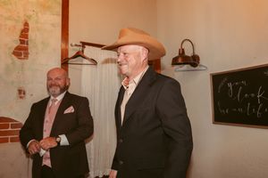 Two men in formal attire stand in a rustic venue, one wearing a cowboy hat.