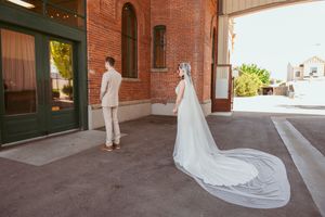 A bride in a long veil stands behind a groom during their first look moment at a brick venue.
