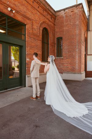 A bride in a flowing veil approaches her groom from behind at a rustic brick venue.