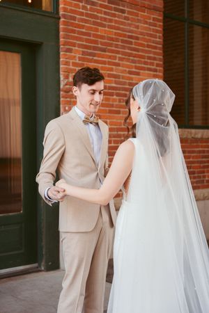 First look wedding photos against a rustic brick wall with bride in white gown and groom in tan suit.