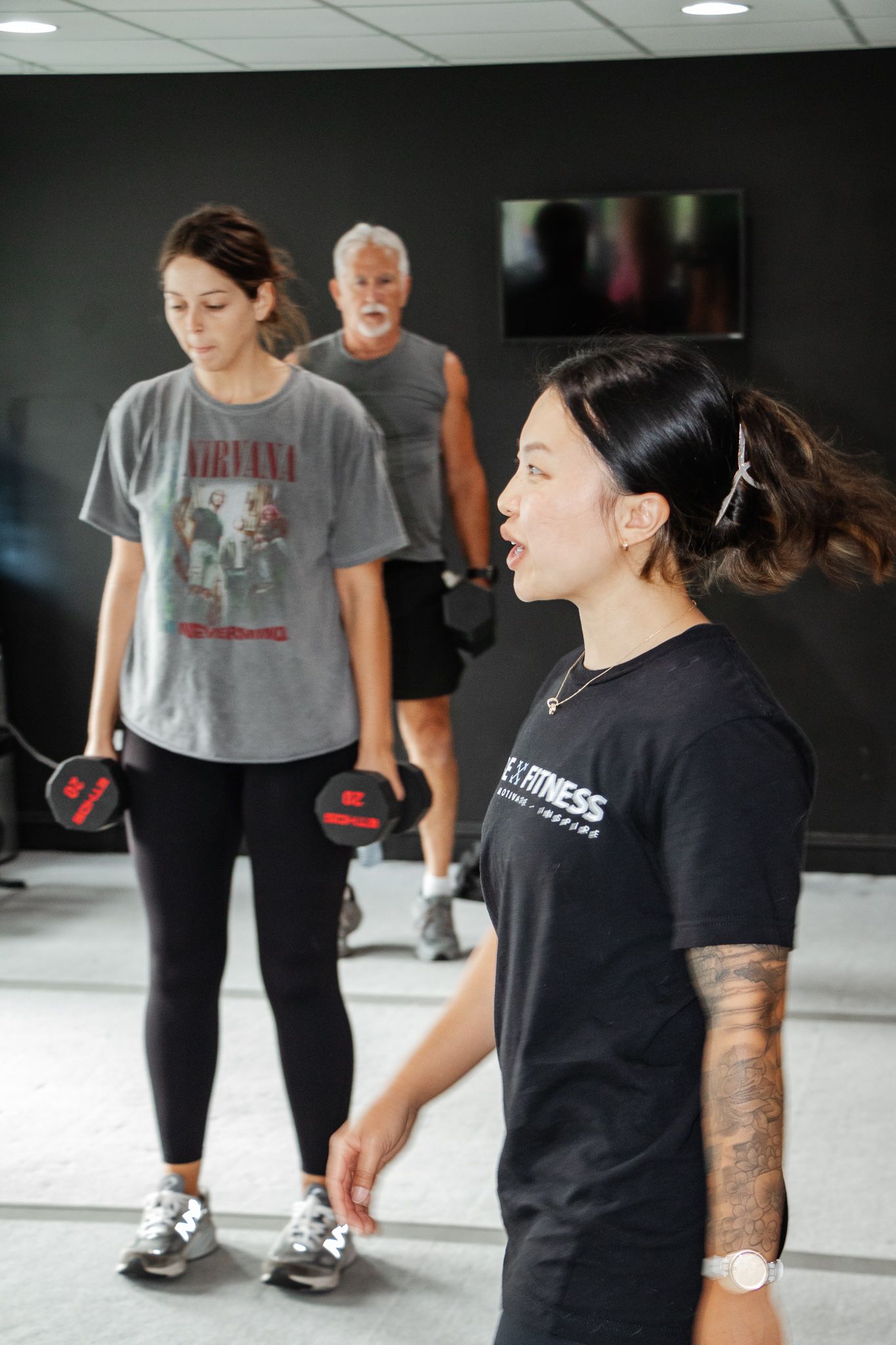 Female fitness instructor in black shirt leading a class, with two participants lifting dumbbells in the background.
