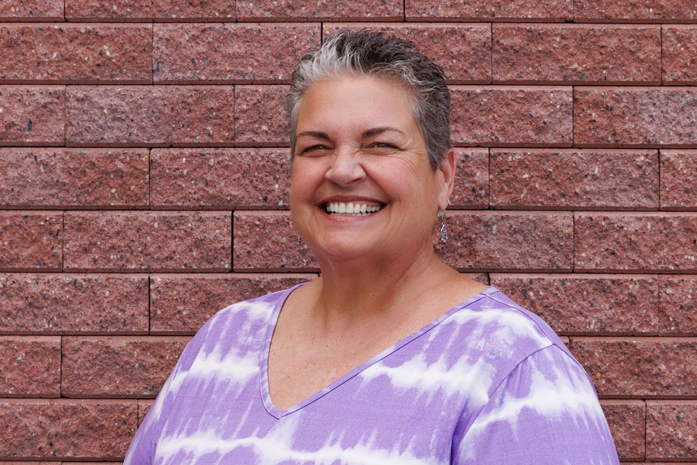 Person wearing purple tie-dye v-neck shirt smiling warmly against a brick wall background.