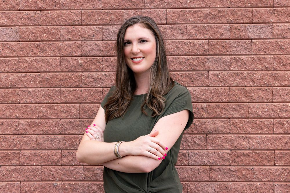 Person in olive green t-shirt standing with crossed arms against a red brick wall.