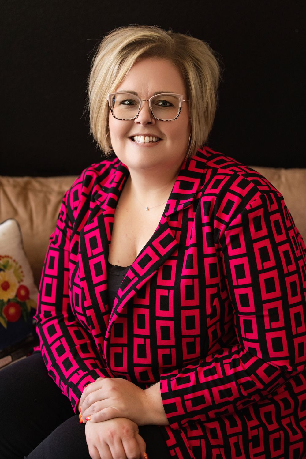 Professional portrait of someone wearing a vibrant pink and black geometric patterned blazer and glasses against dark background.