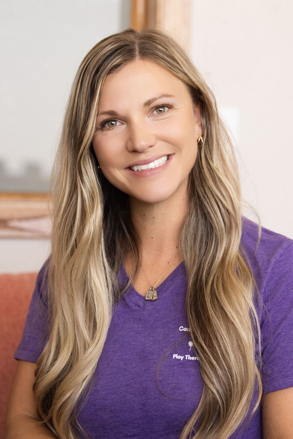 Professional headshot of a person with long blonde wavy hair wearing a purple t-shirt and smiling warmly at the camera.