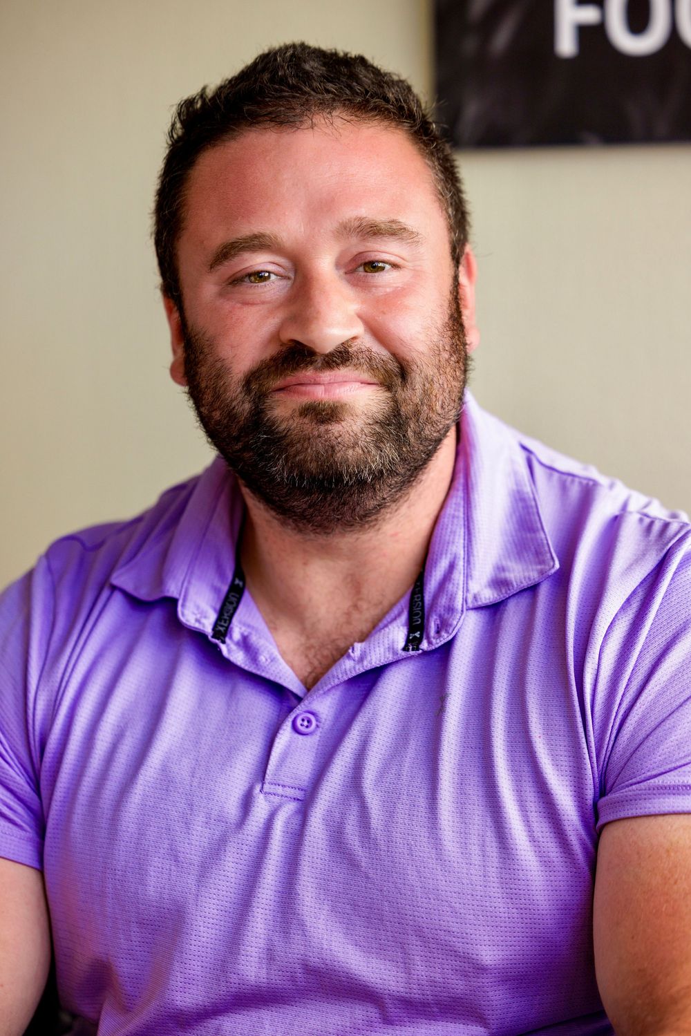 A person in a purple polo shirt smiles warmly while seated indoors with a beard and full cheeks.