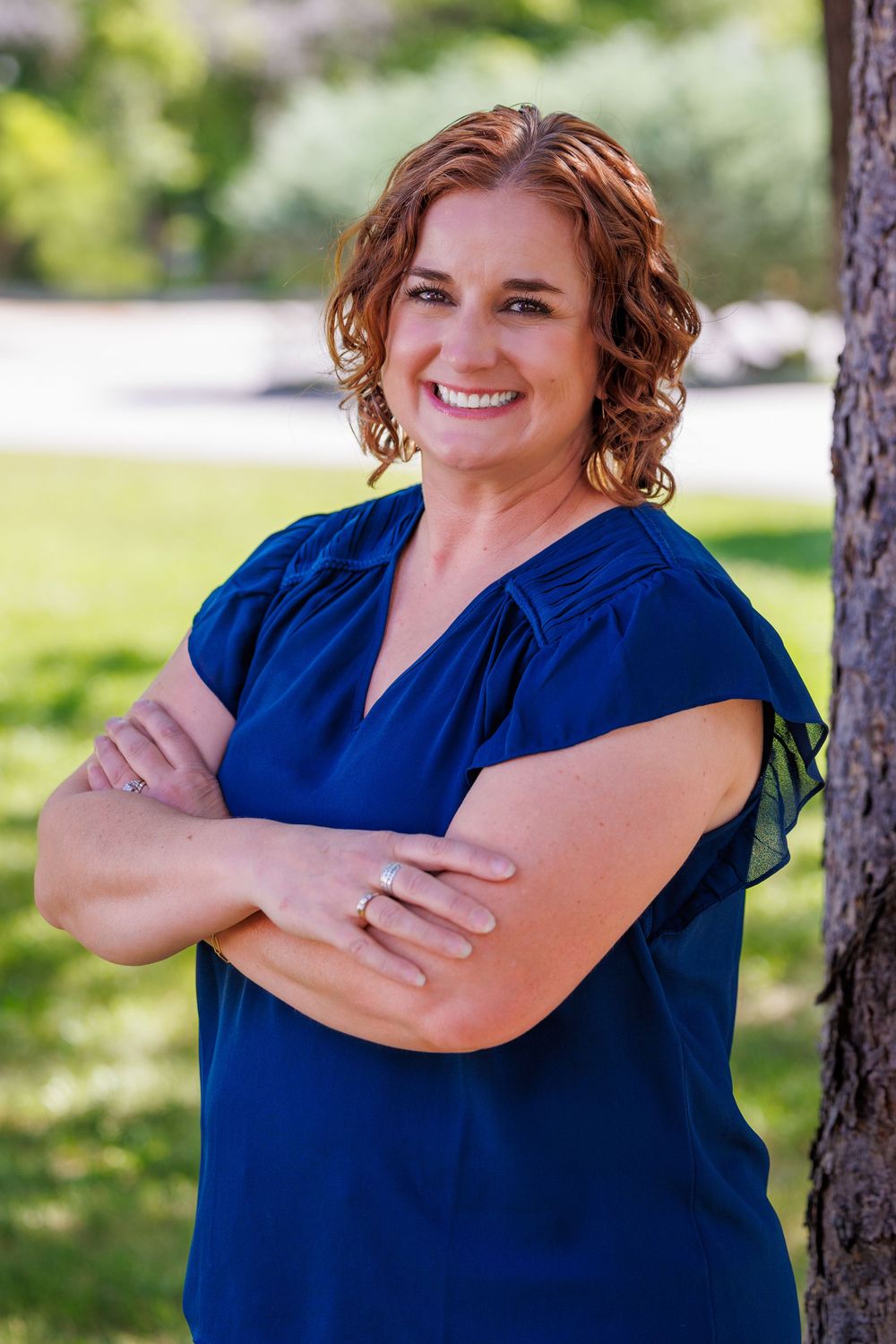 A person in a navy blue short-sleeved blouse poses with crossed arms and a warm smile outdoors near a tree.