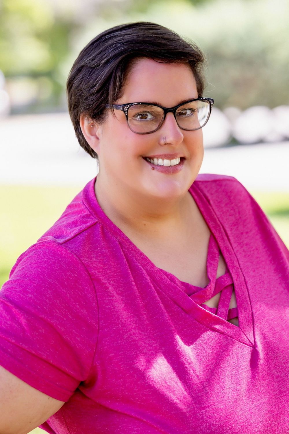 A person wearing glasses and a bright pink polo shirt smiles warmly in an outdoor setting.