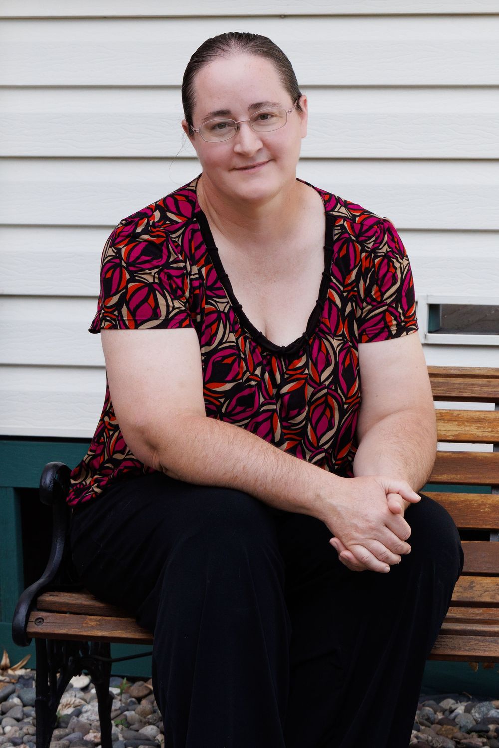 Person in black and red floral print top sits on wooden bench against white siding.