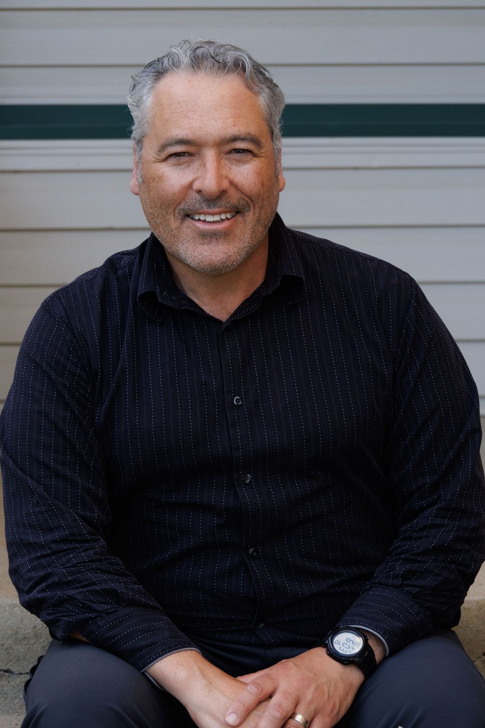 A person seated on outdoor steps wearing a navy pinstripe shirt with rolled sleeves and casual pose against white siding.