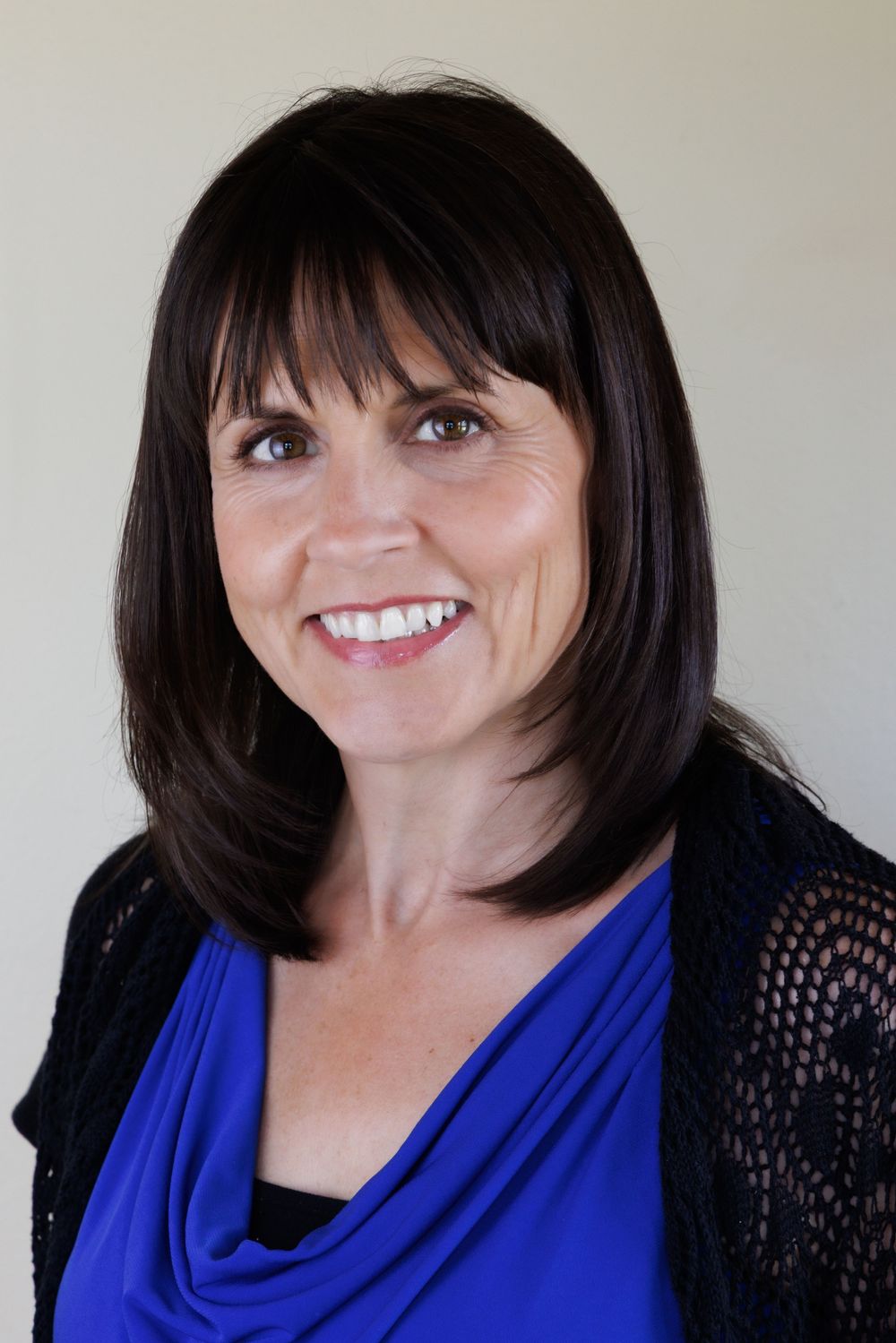 Professional headshot portrait with bright blue top and black lace cardigan against neutral background.