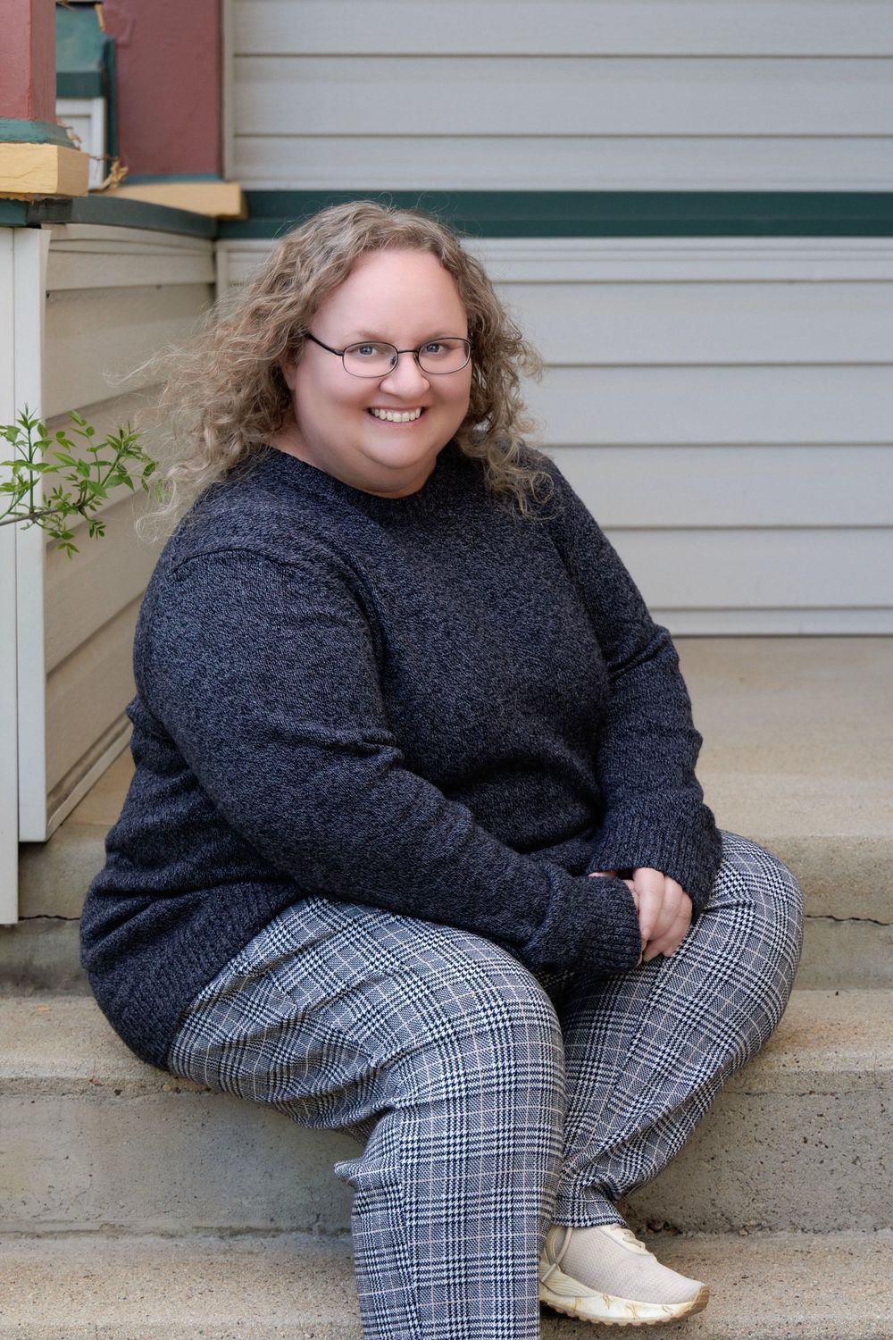 A person in a dark sweater and plaid pants sits smiling on concrete steps next to greenery.