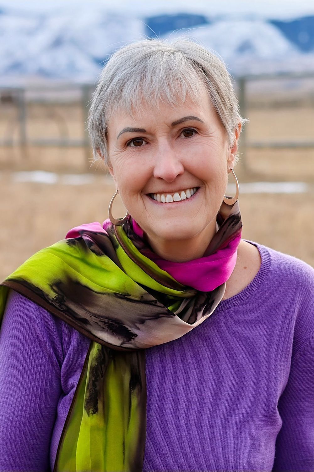 Woman wearing purple sweater and colorful patterned scarf poses outdoors with winter mountains in background.