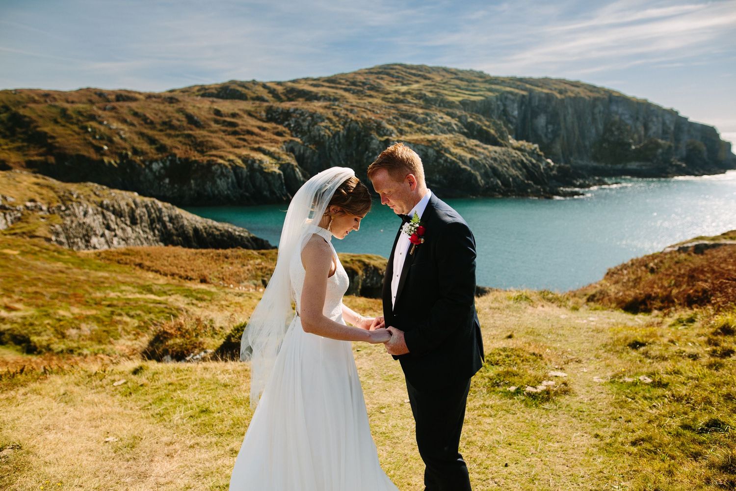 A couple shares a romantic moment on coastal cliffs overlooking a blue ocean bay on their wedding day.