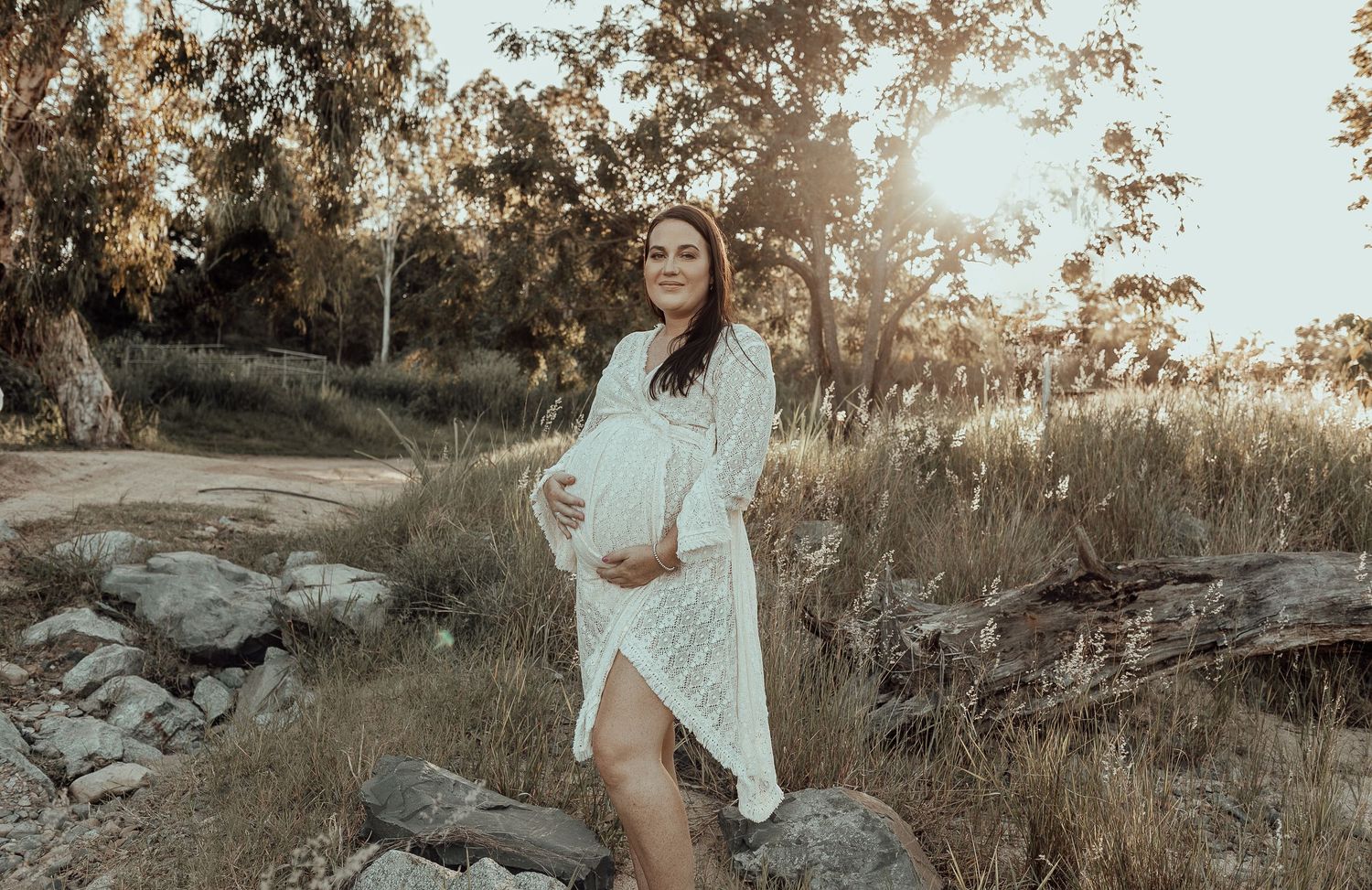 Expectant mother in white dress poses on rocks at sunset in a natural outdoor setting.