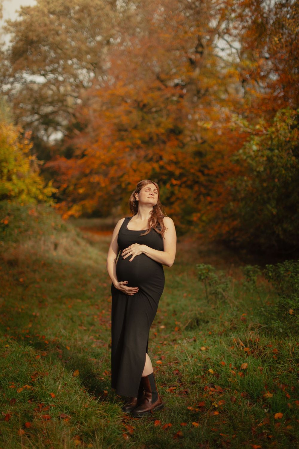 A pregnant woman in a black dress poses outdoors during autumn with vibrant orange and yellow foliage in the background.