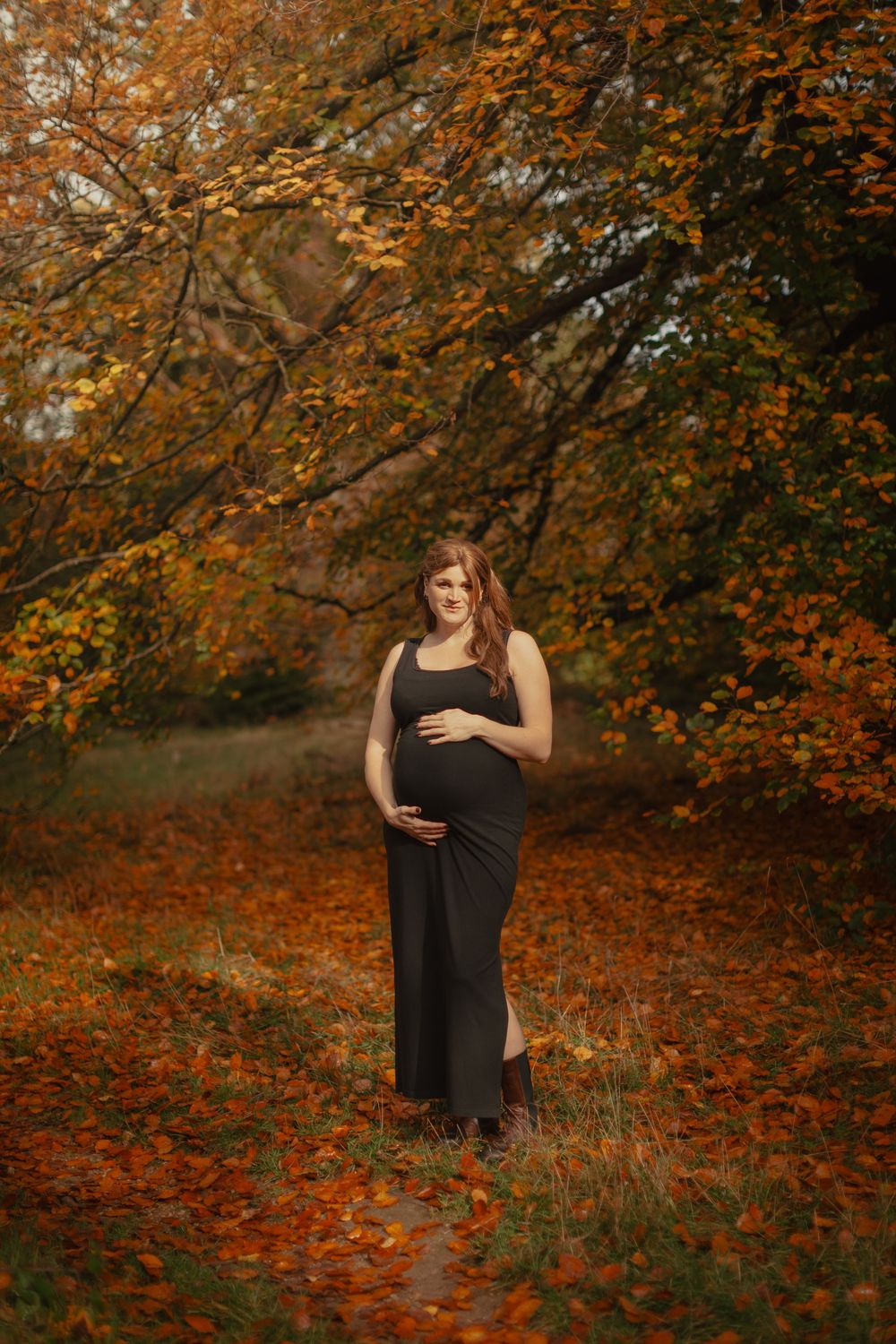 An expectant person in a long black dress stands among autumn foliage with orange and brown leaves.