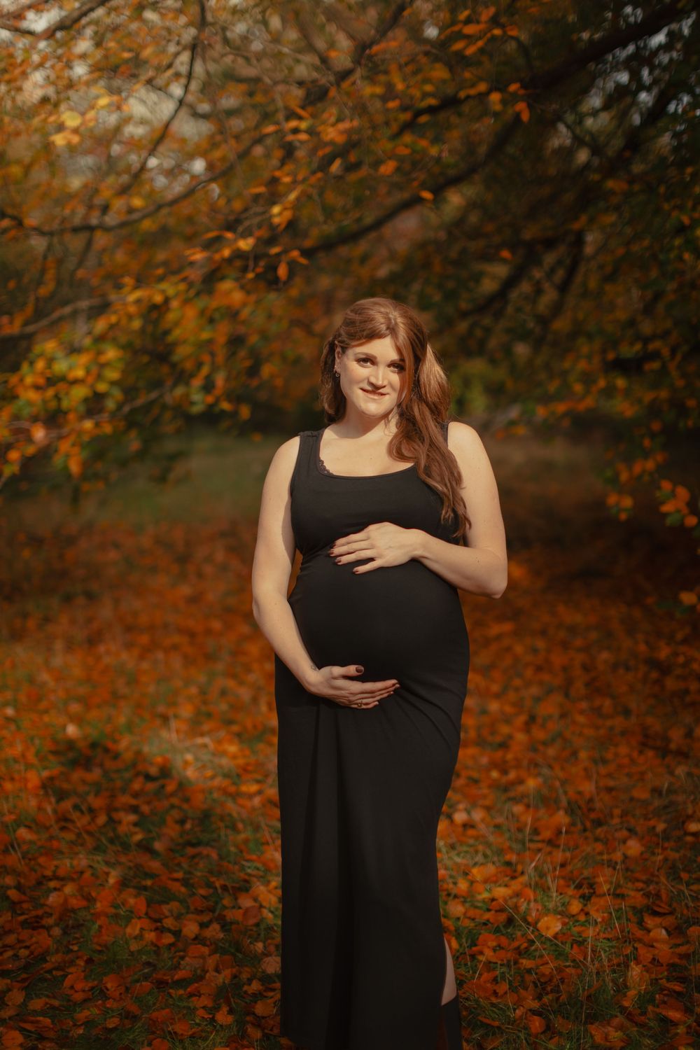 A pregnant woman in a black dress poses gracefully against an autumn forest backdrop with orange and brown foliage.