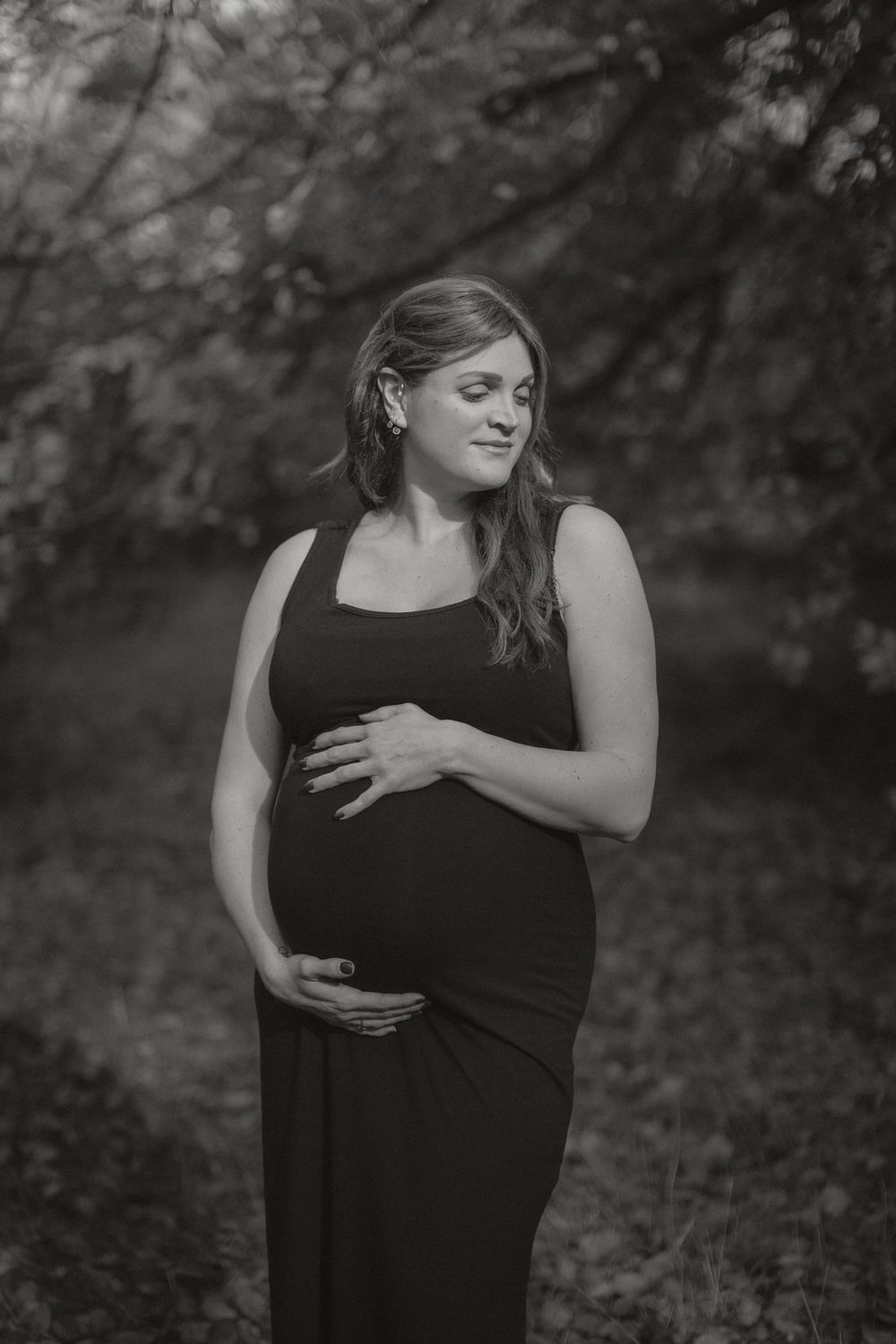 A black and white maternity portrait showcasing an expectant mother in a black dress against a natural outdoor backdrop.